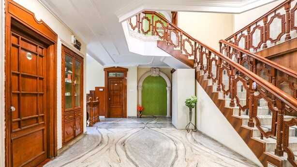 Un couloir au sol en marbre, doté de portes en bois et d’un escalier orné de balustrades travaillées.