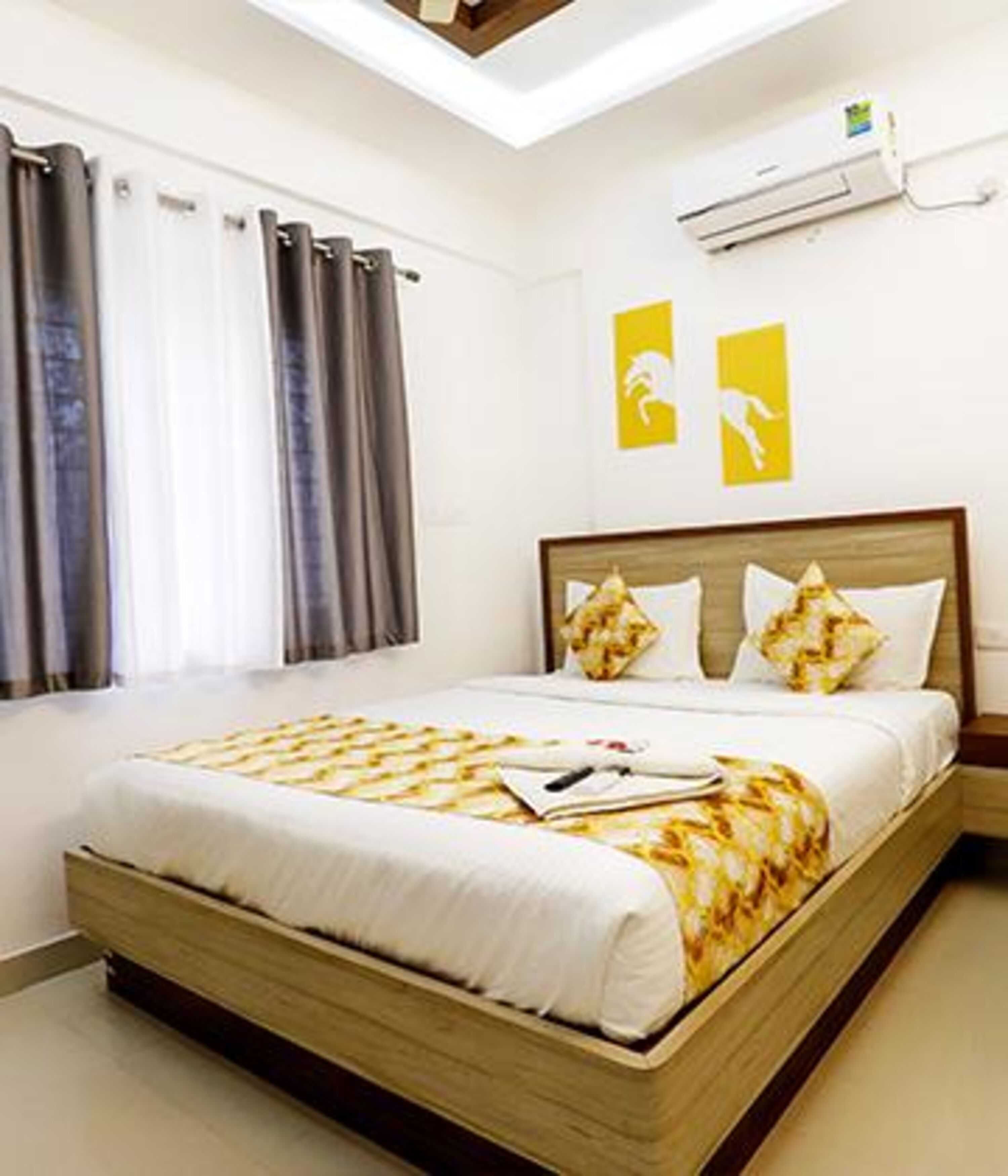 A bedroom with a large bed, a window with curtains, an air conditioning unit, and two yellow wall art pieces.