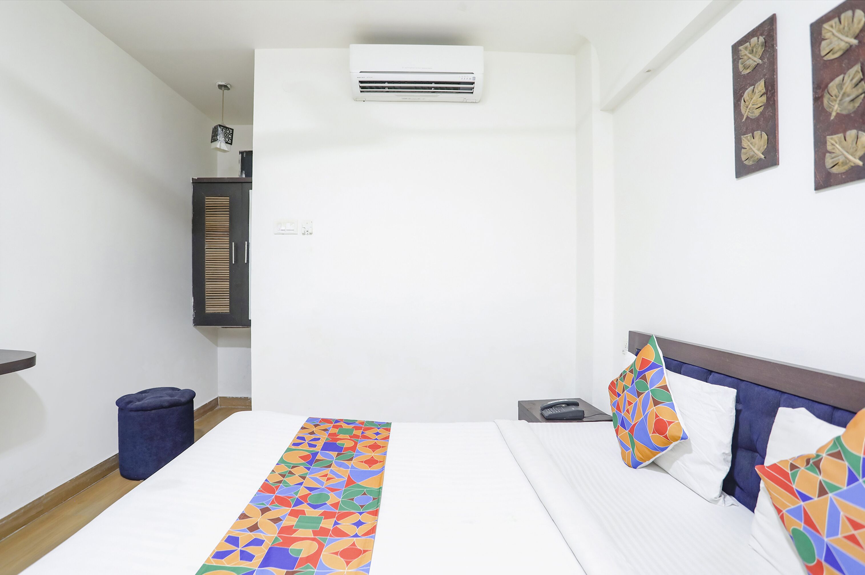 Une chambre d’hôtel comprenant un lit, une petite table de chevet, un appareil de climatisation, une étagère fixée au mur et un coussin décoratif.