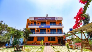 A multi-story building with a sign for 'Prajakta Holiday Inn' and 'Fab Hotels'. It features a central courtyard with a checkered floor pattern, surrounded by potted plants and outdoor seating. - Fabexpress Prajakta Holiday Inn (Mahabaleshwar)