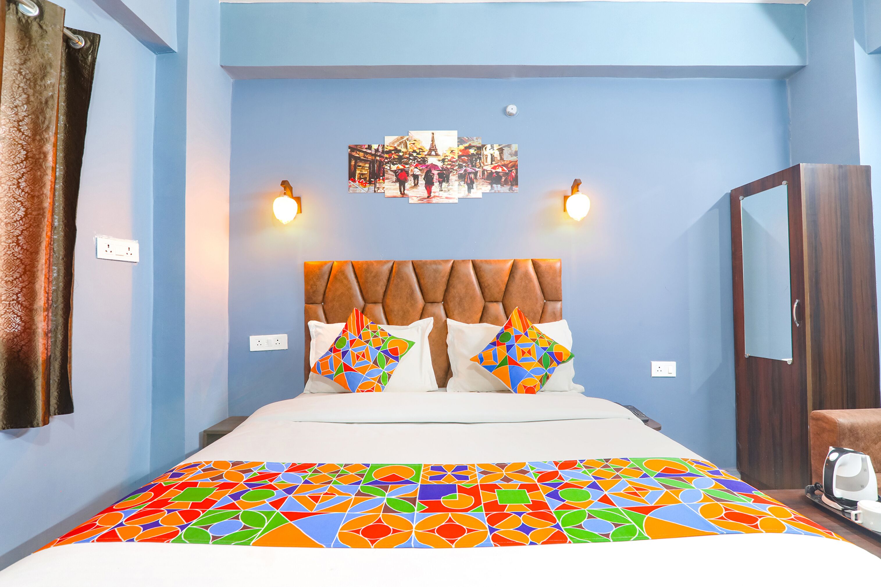 A hotel room with a bed, a headboard, two wall-mounted lights, a wall-mounted picture, and a wardrobe.