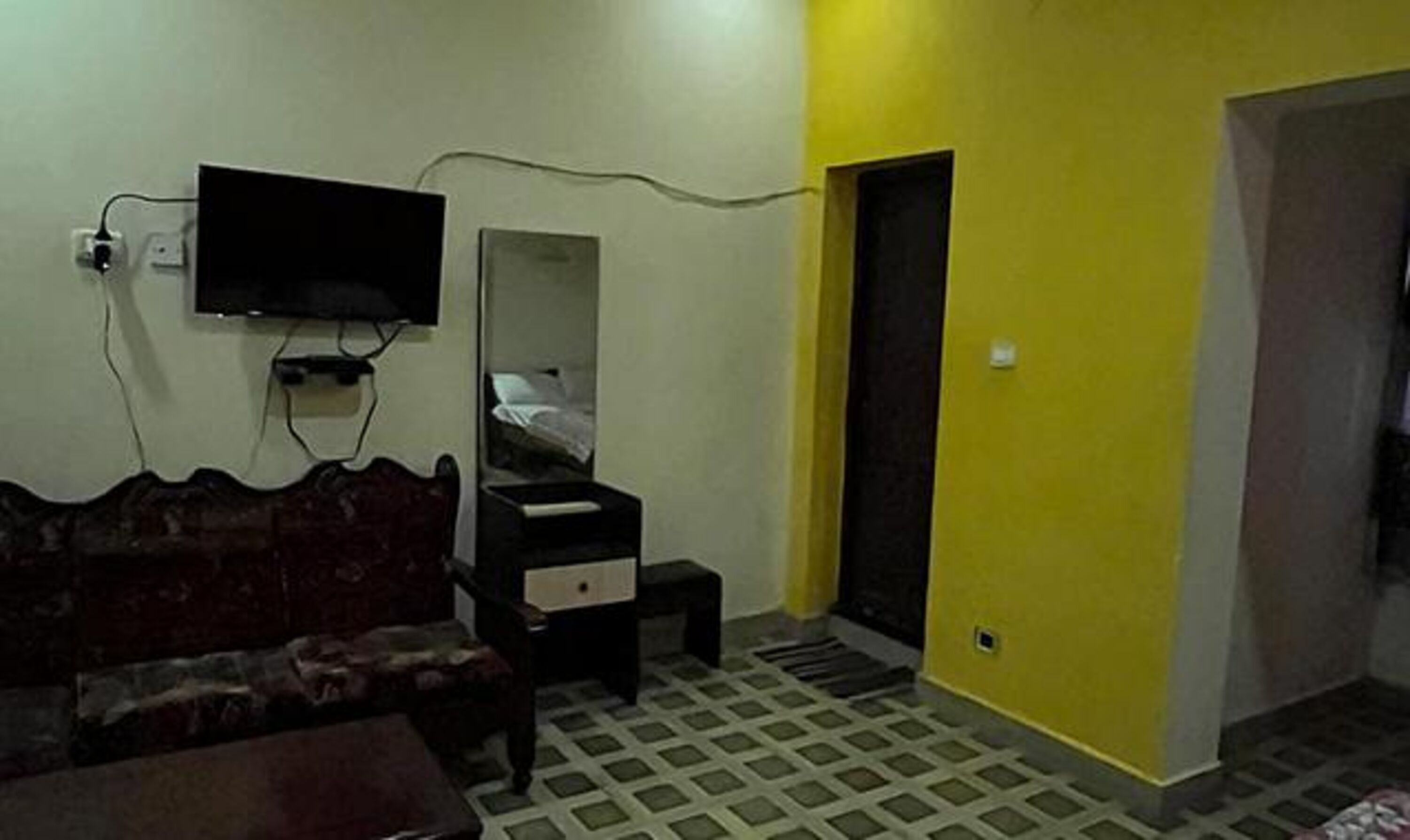 A room with a sofa, a small table, a TV mounted on the wall, a mirror, and a door leading to another room.