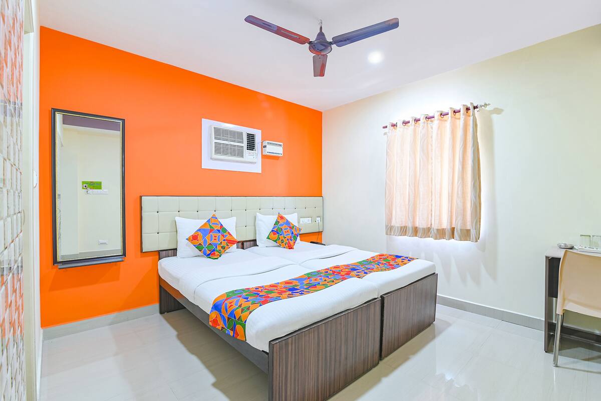 Een hotelkamer met een oranje muur, een bed met een kleurrijke deken, een plafondventilator, een raam met gordijnen en een klein tafeltje met een stoel.