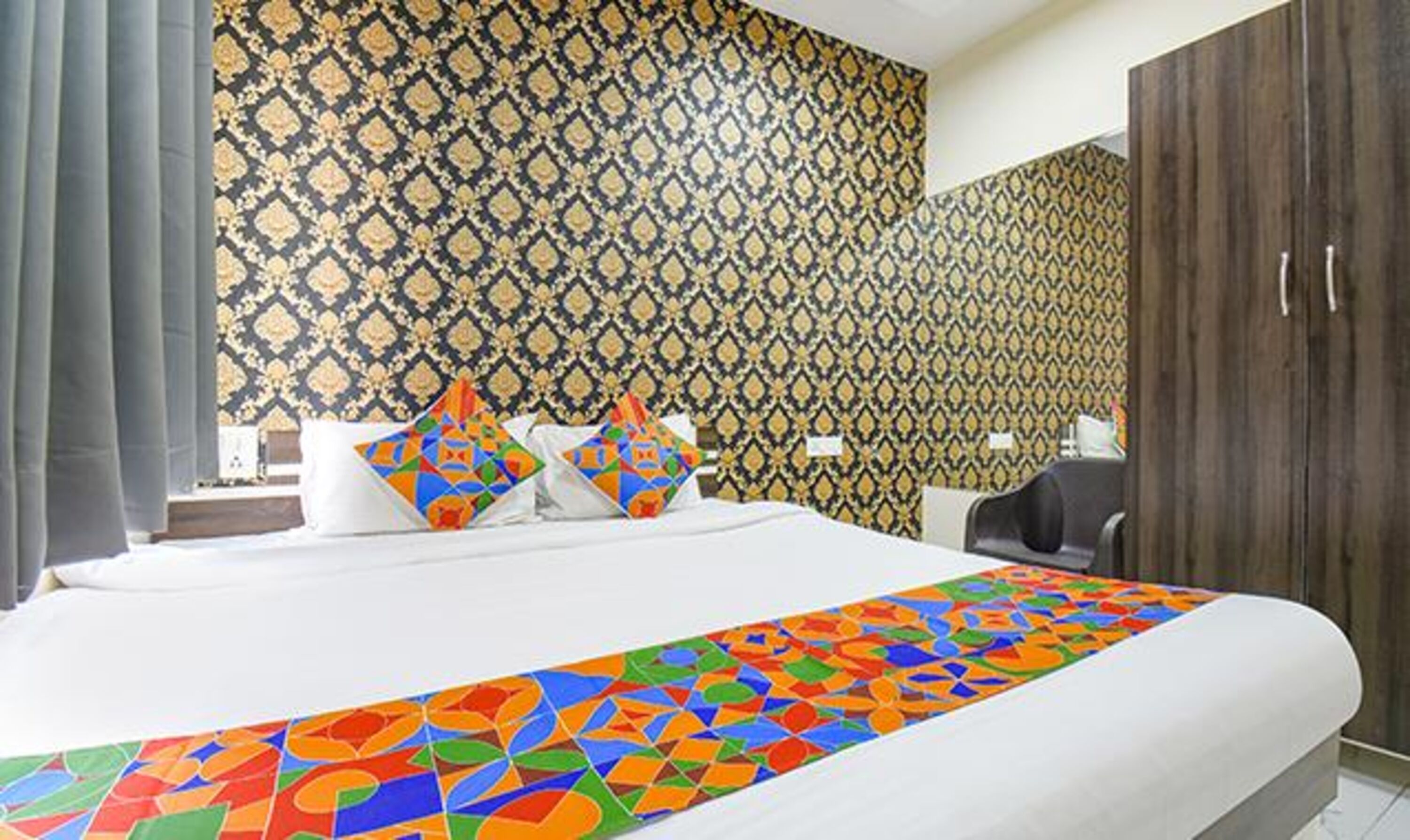 A hotel room with a patterned wallpaper, a bed with colorful pillows, and a wooden wardrobe.