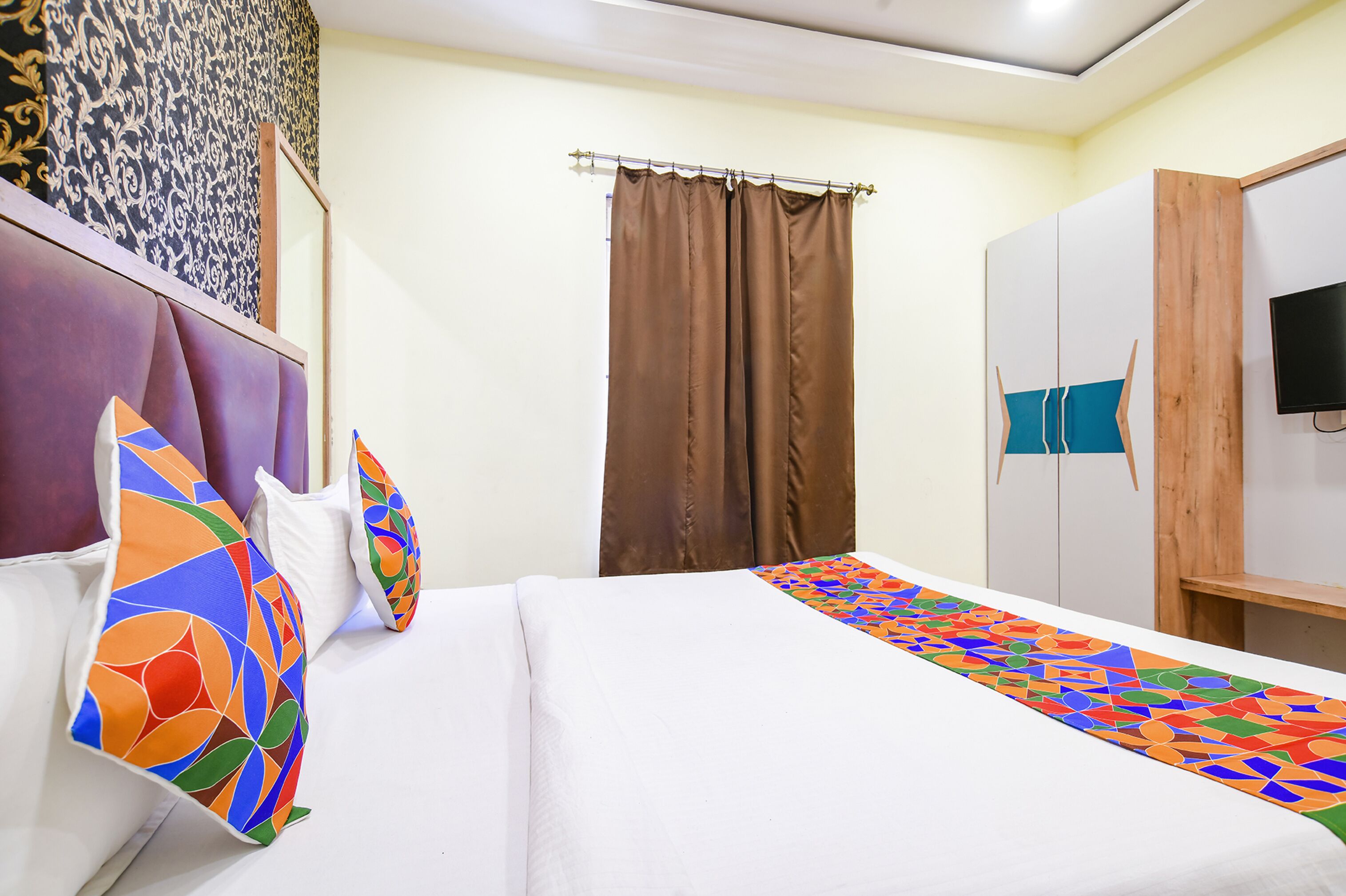 Una cama bien hecha con almohadas coloridas, un armario de madera y un televisor en la pared.