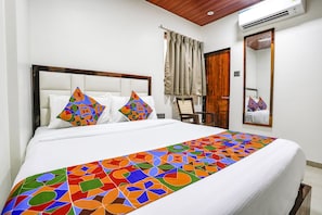 A neatly made bed with colorful geometric-patterned bedspread and pillows, a wooden headboard, a wooden chair, a mirror, and a door with a wooden frame.