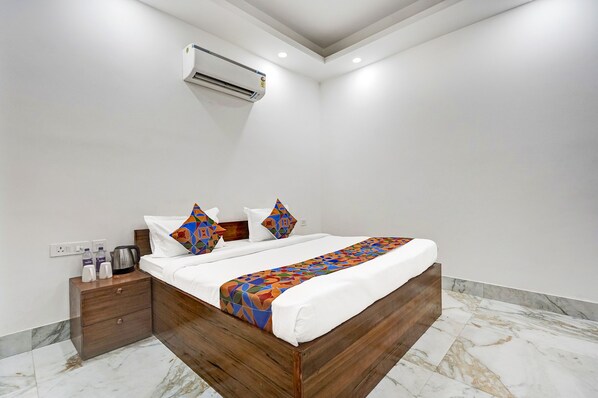 Quarto de hotel moderno com cama de madeira, roupa de cama branca e manta colorida. Há uma mesa de cabeceira com chaleira e tomada.