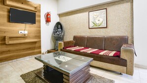 Ein Wohnzimmer mit Flachbildfernseher an einer Holzwand, einem braunen Ledersofa mit roter und weißer Decke, einem Glastisch und einer Buddha-Statue aus Stein.