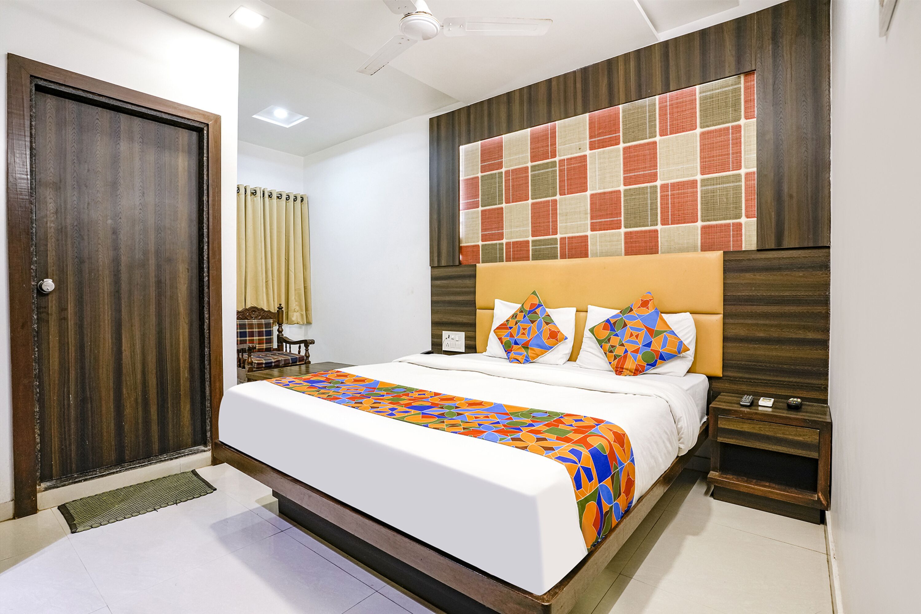 A neatly made bed with a colorful geometric-patterned bedspread, a wooden headboard, and a bedside table with a remote control.
