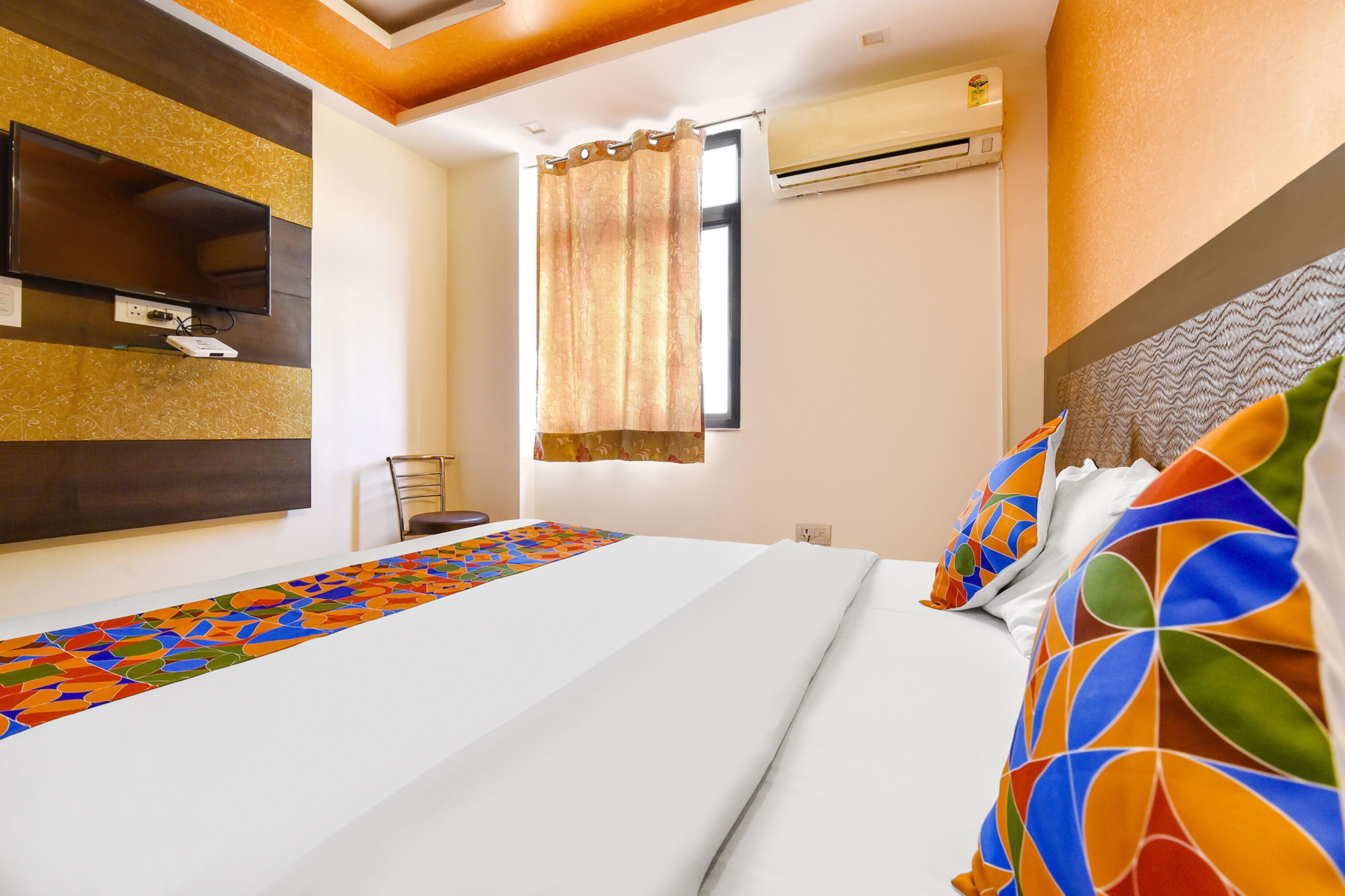 A hotel room with a bed, a TV mounted on the wall, an air conditioning unit, and a window with curtains.