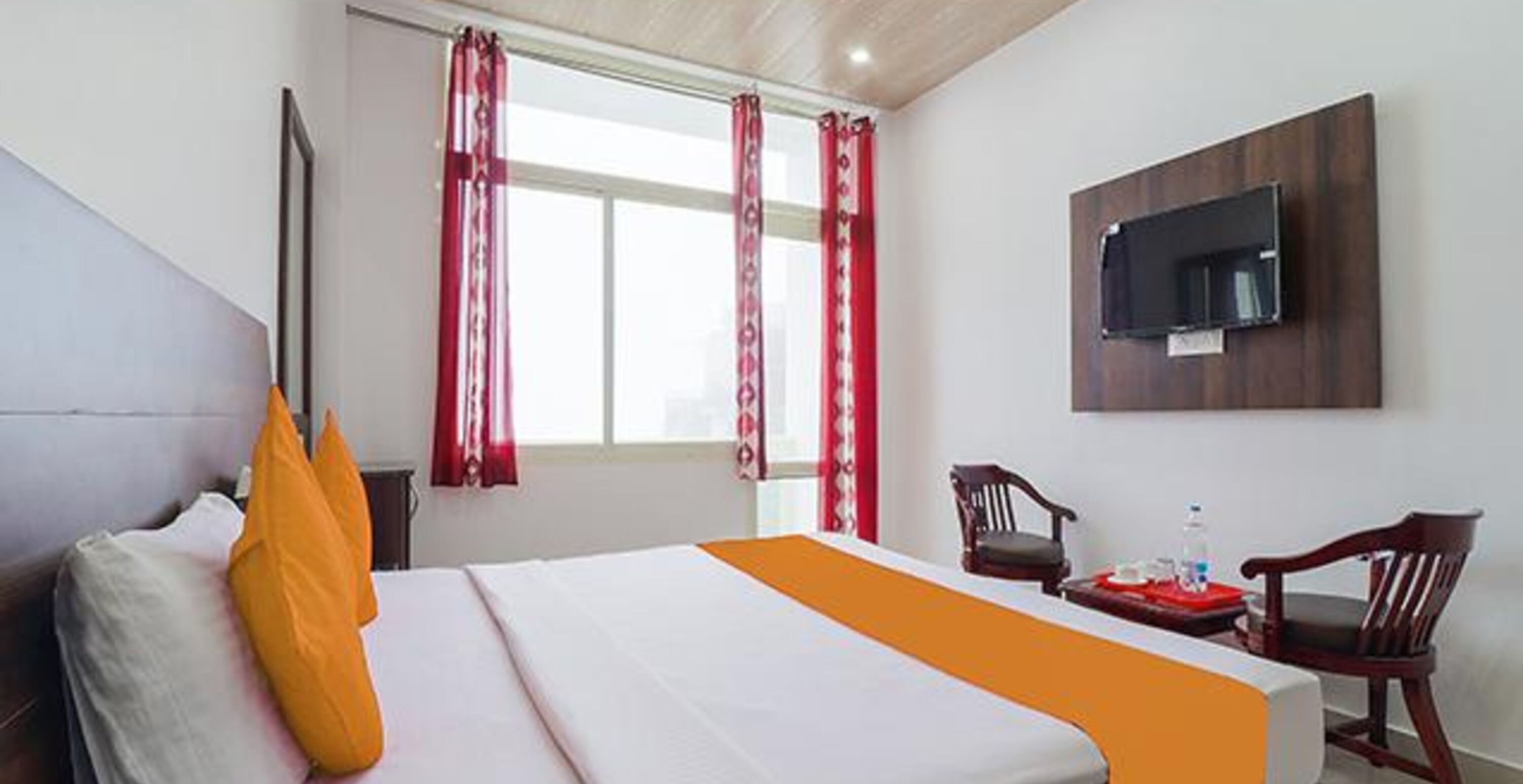Ett hotellrum med en säng, en tv som är monterad på väggen, ett skrivbord med en stol och ett fönster med gardiner.