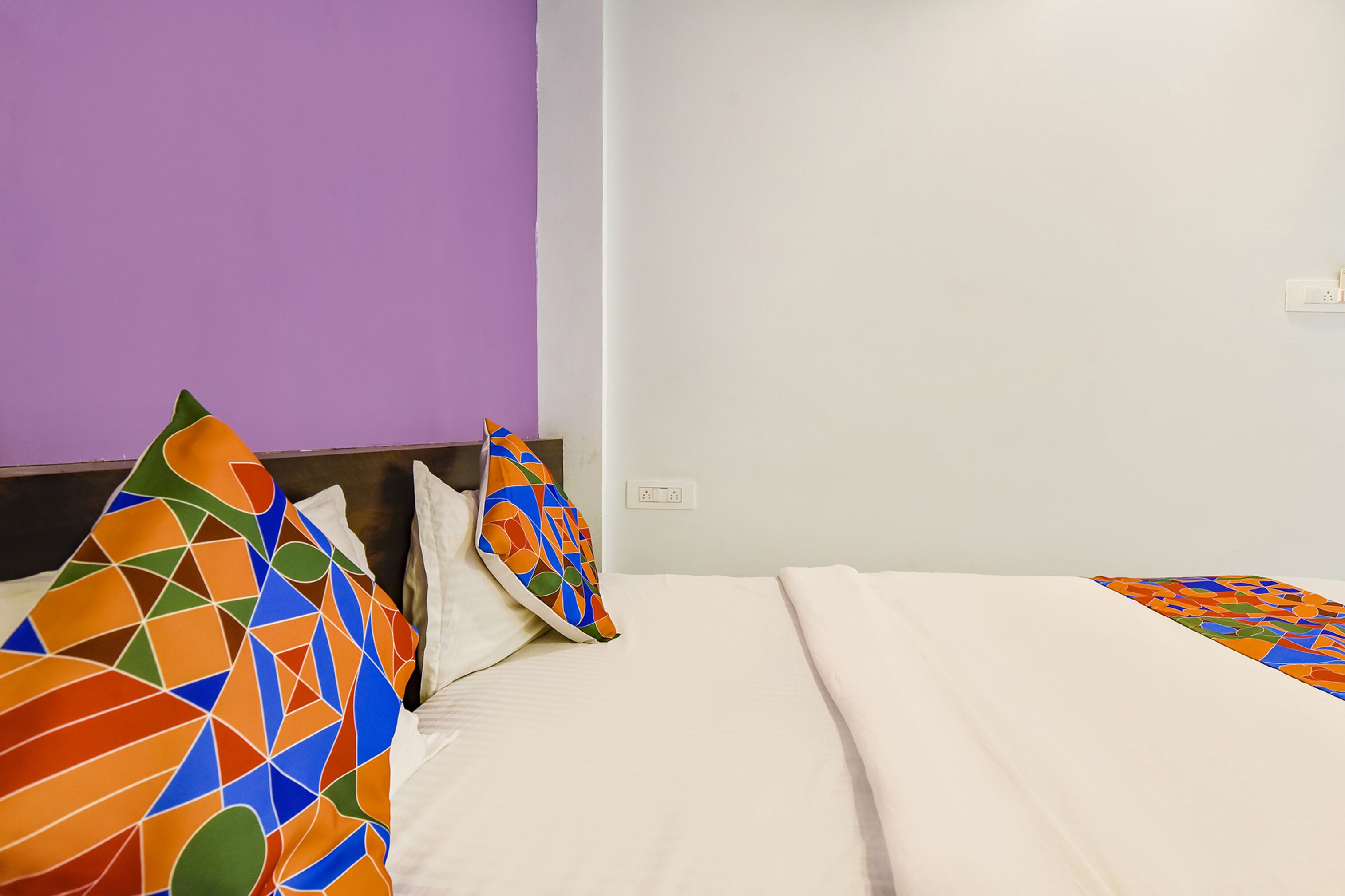 A hotel room with a purple wall, a white bed, and colorful geometric-patterned pillows.