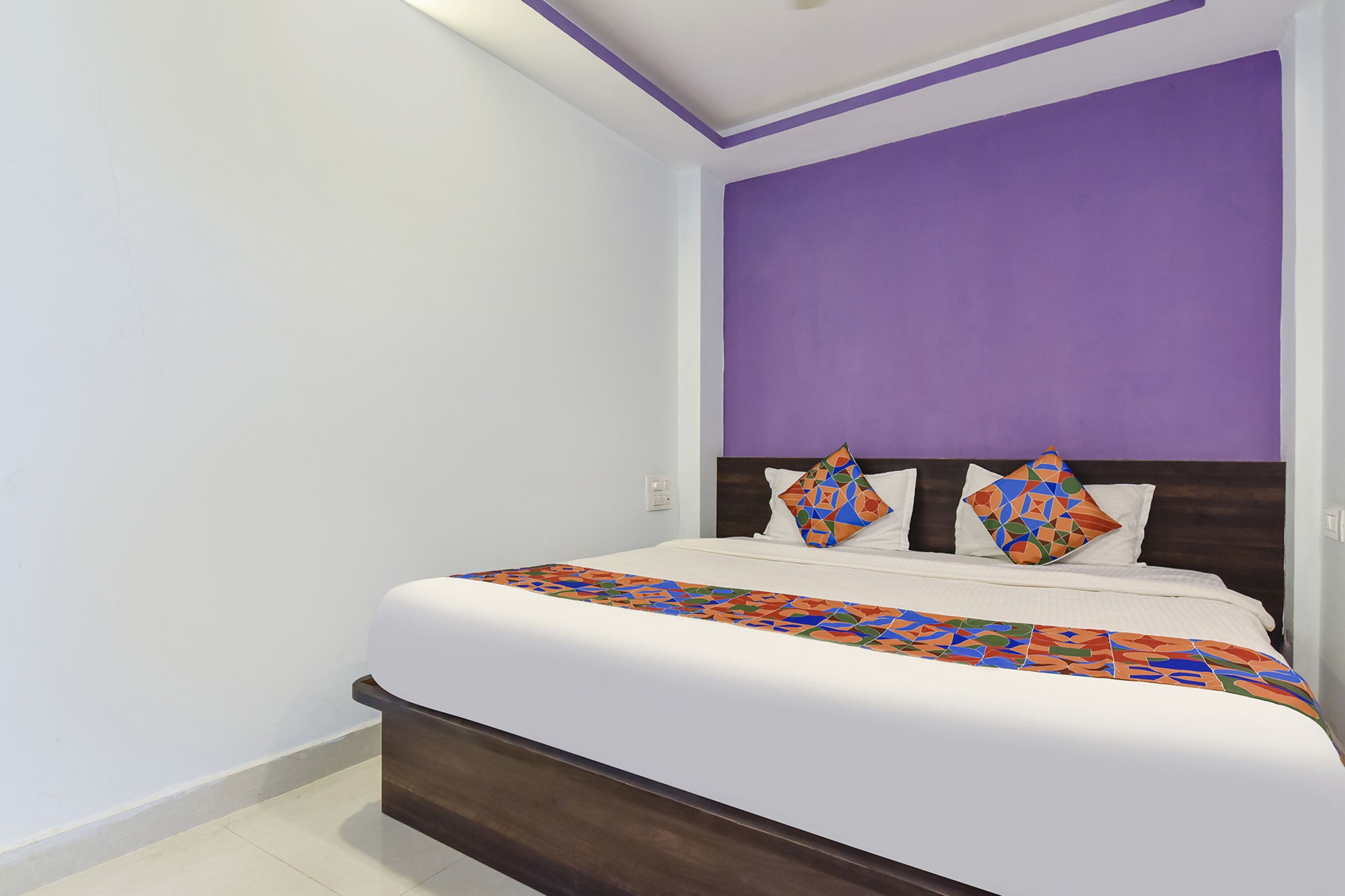 Una cama bien hecha con una colcha de diseño geométrico y colores vivos, un cabecero de madera oscura y una pared de acento color púrpura.
