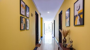 A hallway with yellow walls, framed artwork, a wooden console table with books and a vase, and a door at the end. - FabHotel Arambol Inn (Arambol)