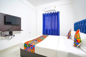 A hotel room with a bed, a TV mounted on the wall, an air conditioning unit, and a window with blue curtains.
