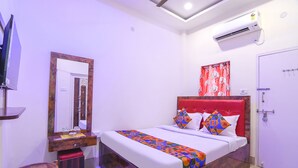 A hotel room with a bed, a mirror, a vanity with a mirror, a red headboard, a white door, and an air conditioning unit. - Fabexpress Vedika (Kolkata)