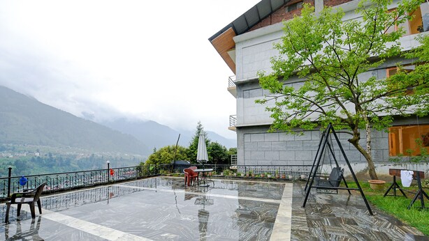 A terrace with a view of mountains, a swing, and a table with chairs. - Fabhotel Attithi Huts And Cottage (Manali)