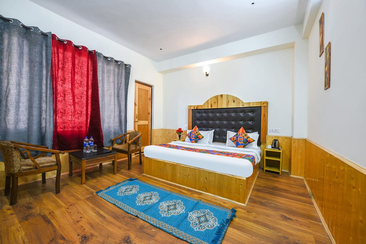 A bedroom with a wooden headboard, a bed with colorful pillows, a blue patterned rug, two wooden chairs, a small table with water bottles, and a wooden door.