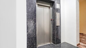 Un ascensor con acabado metálico, ubicado en un pasillo con paredes de mármol y piso de baldosas.