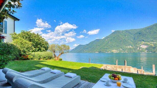 A deck with lounge chairs and a table set for breakfast overlooking a lake and mountains. - Maison Sur l'Eau - Lugano Villa with View Terrace, Paradiso (Lugano), Switzerland (Paradiso (Lugano))