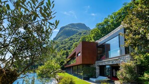 A modern house with a stone facade, large windows, and a view of a lake and mountain. - Maison Sur l'Eau - Lugano Villa with View Terrace, Paradiso (Lugano), Switzerland (Paradiso (Lugano))