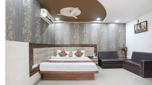 A modern hotel room with a large bed, a wooden headboard, a dark leather sofa, a small table with a vase of flowers, and a ceiling fan. - Fabhotel Royal City (Ahmedabad)