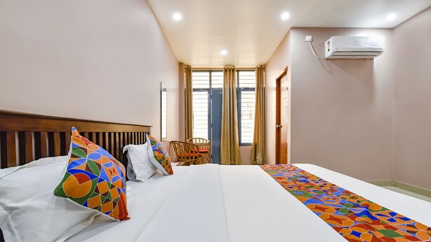 Habitación de hotel con una cama grande, cabecera de madera, almohada colorida, una silla y una ventana con cortinas.