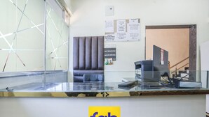 Una recepción de hotel con un separador de vidrio, una computadora y un área de descanso con un sofá. Hay carteles en la pared con información sobre tarifas y políticas de habitaciones.
