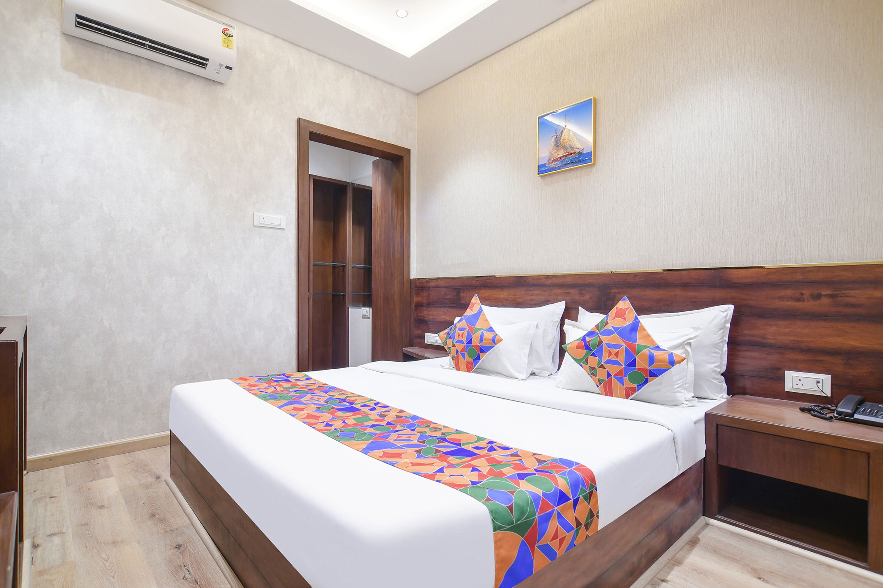 A hotel room with a large bed, wooden headboard, and a colorful bedspread. There is a small bedside table with a phone and a nightstand with a lamp.