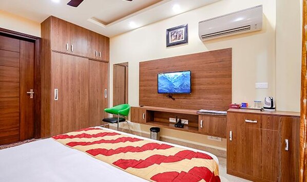A hotel room with a large bed, wooden wardrobe, a flat-screen TV mounted on the wall, a small desk with a chair, and an air conditioning unit.