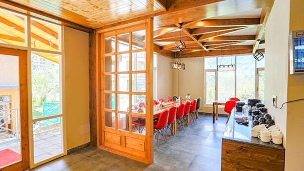 A spacious room with a large wooden door leading to an outdoor area, a long table with red chairs, and a shoe rack on the right. - Fabhotel Outhills (Manali)