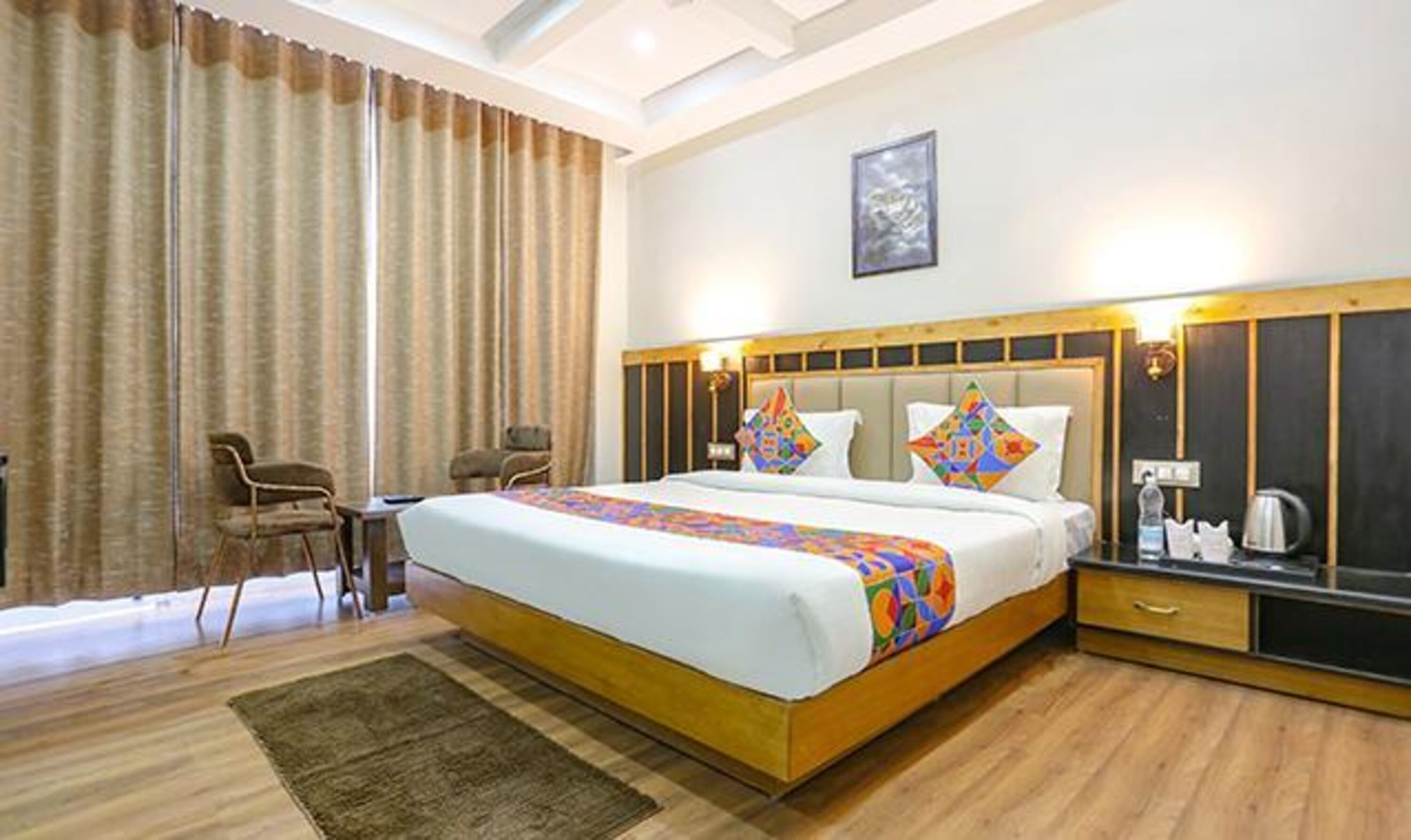 A hotel room with a large bed, two chairs, a small table, and a nightstand with a kettle.