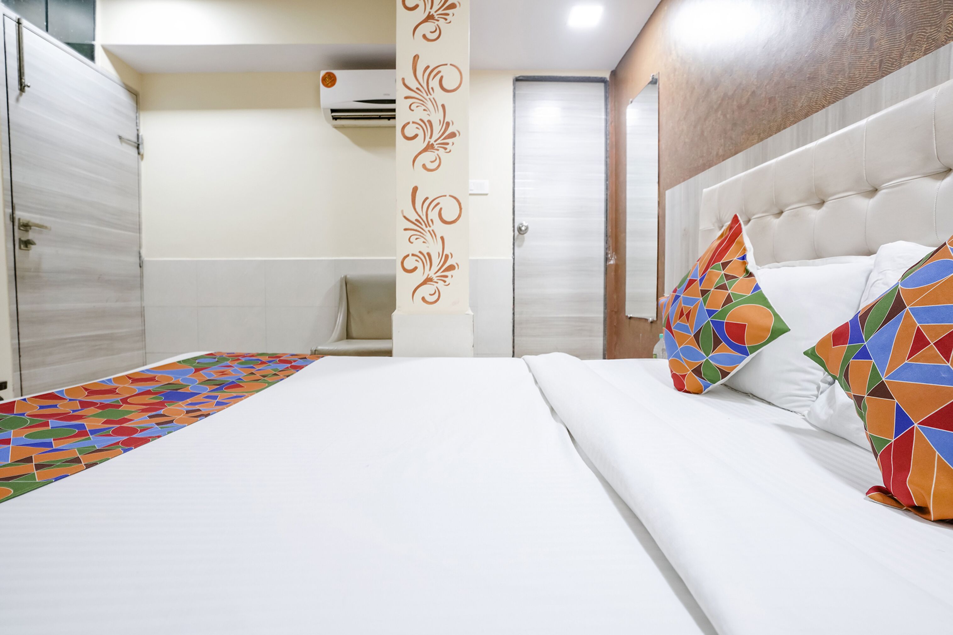 A hotel room with a bed, colorful pillows, a decorative wall panel, and a bathroom with a shower and toilet.