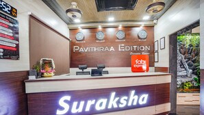 A reception desk with a sign reading "Suraksha" and "Pavithraa Edition" above it. There are clocks showing different time zones, a digital clock, and a "Fab Express" sign. - FabHotel Pavithraa Edition (Bengaluru)