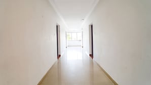 A long, narrow hallway with white walls and a shiny tiled floor, leading to a bright room with a large window. - FabExpress California Inn (Kodaikanal)