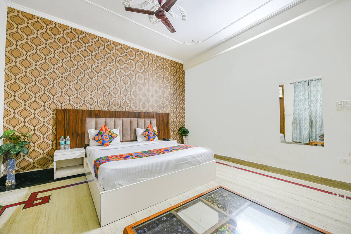 Una habitación de hotel con papel tapiz estampado, cabecera de madera, cama con colcha colorida, mesita de noche con planta y una mesa de centro de cristal.