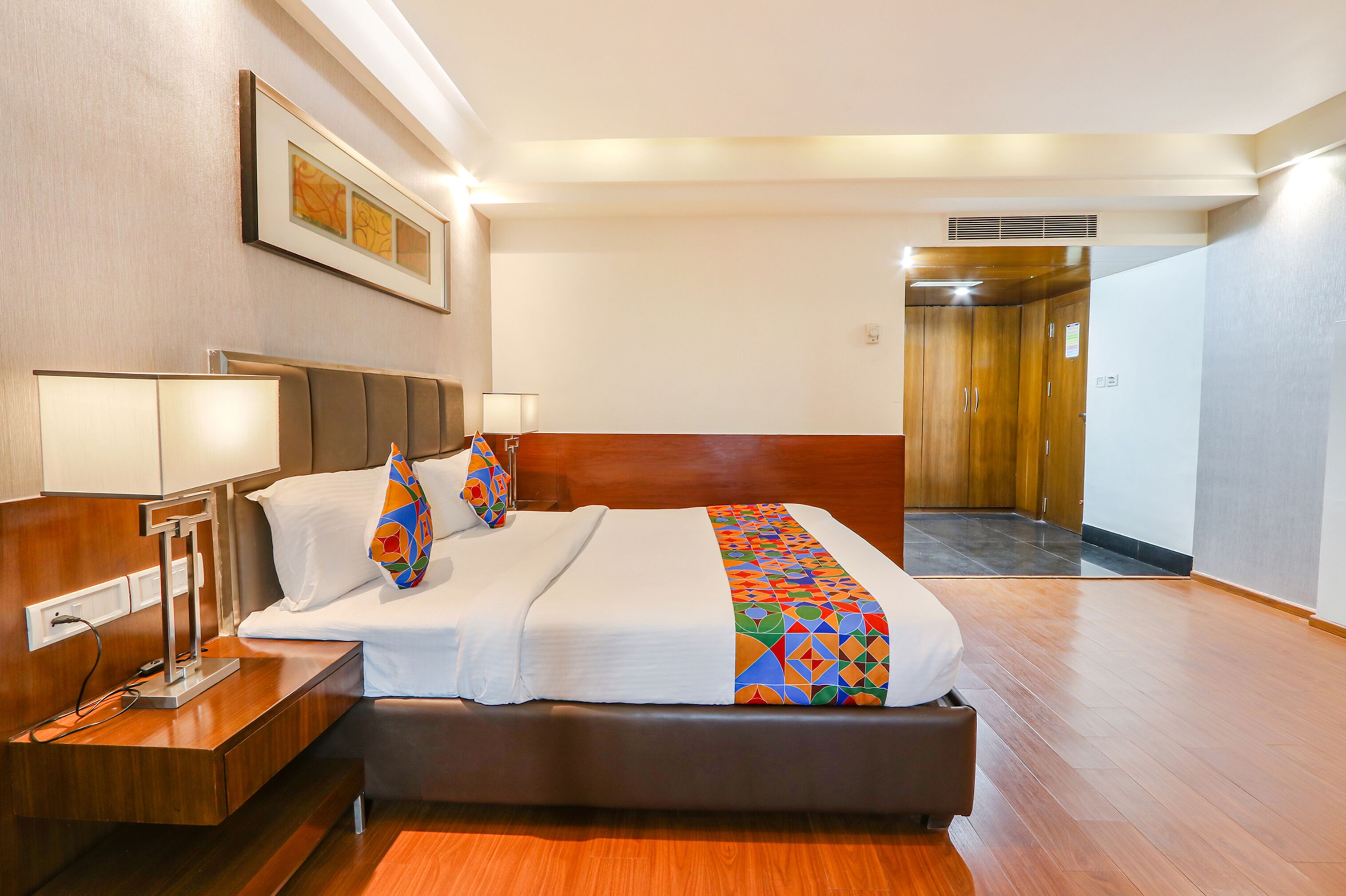 A hotel room with a bed, bedside tables, a lamp, a wall-mounted artwork, and a wooden wardrobe.