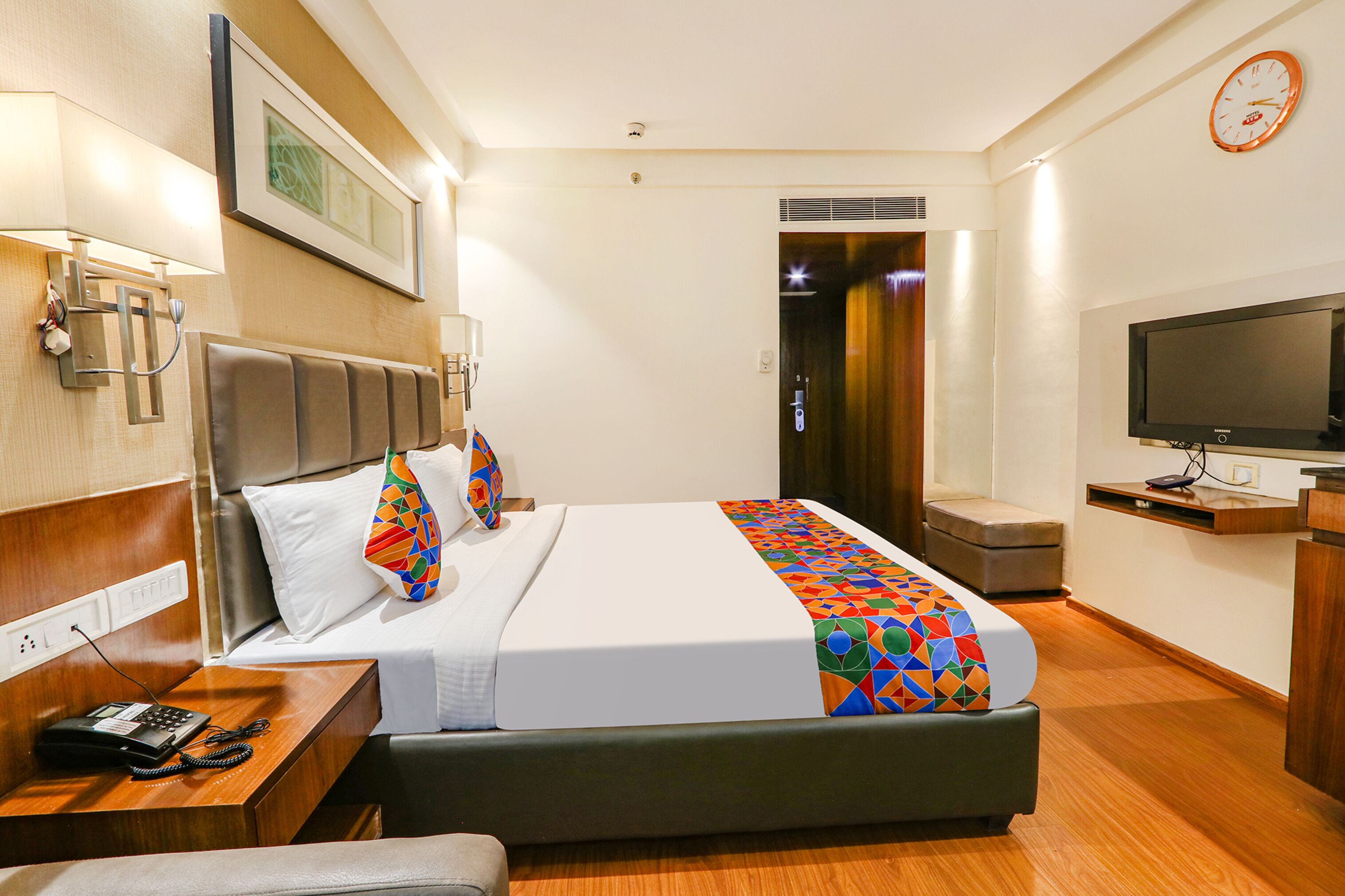 Una habitación de hotel con cama, mesita de noche, televisor y un reloj en la pared.