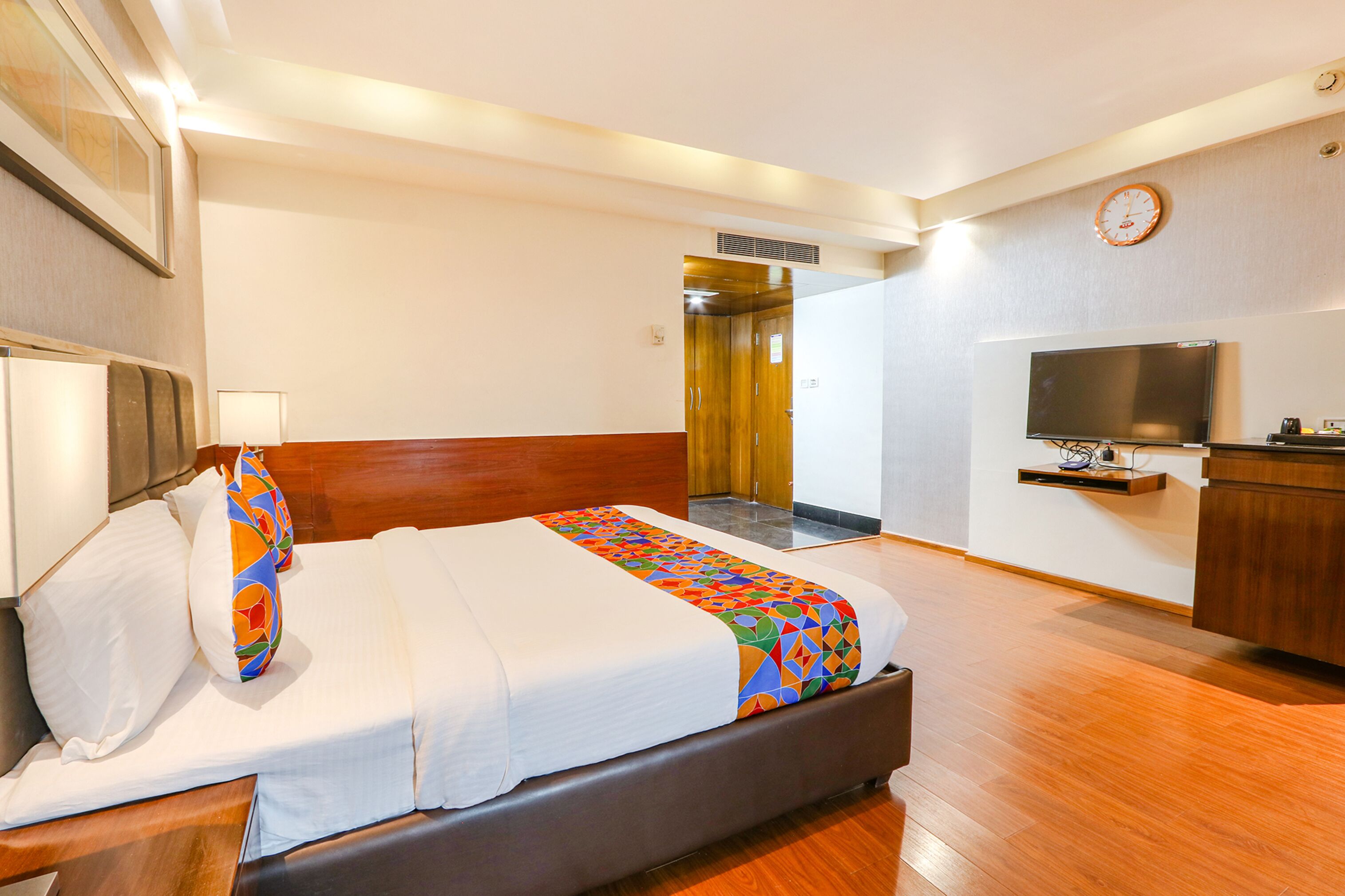 A hotel room with a bed, a television, a desk, and a clock on the wall.