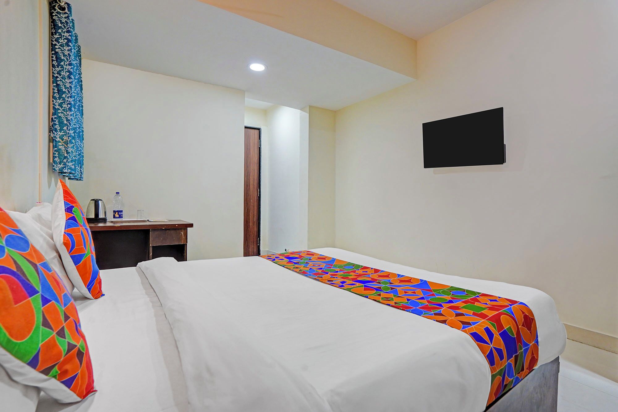 A hotel room with a bed, a desk, a chair, a water bottle, a kettle, and a TV.