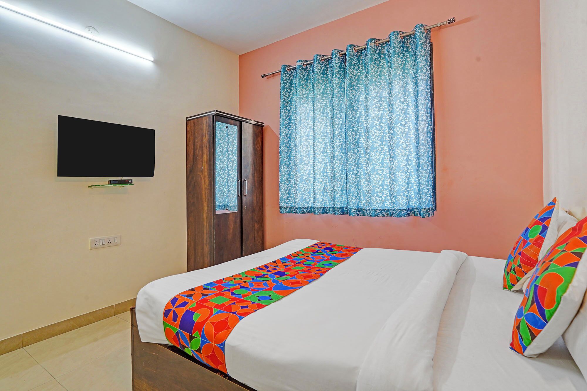 A hotel room with a bed, a television mounted on the wall, a wardrobe, and a window with curtains.