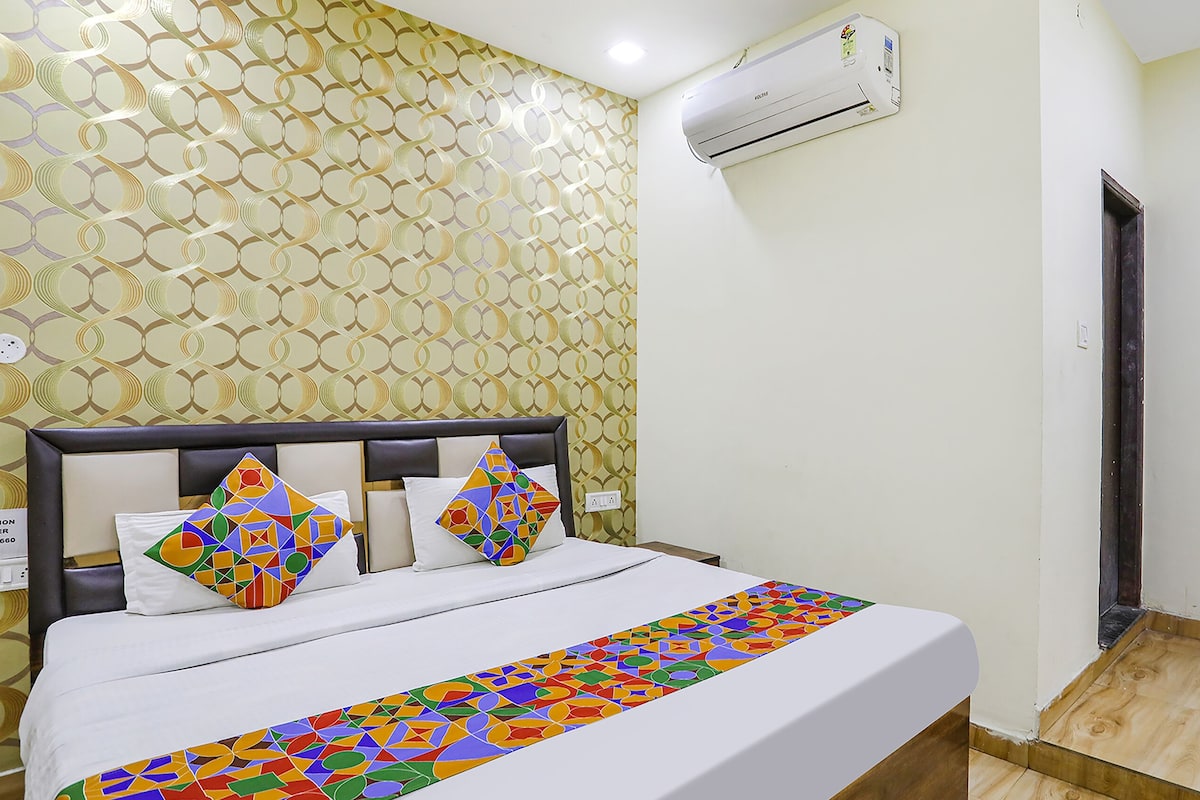 A hotel room with a patterned wallpaper, a bed with colorful pillows, a white bedside table, and an air conditioning unit on the wall.