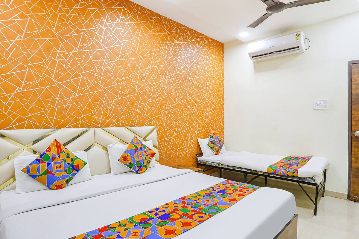 A hotel room with a geometric patterned wall, a bed with colorful pillows, a bedside table, and an air conditioning unit.