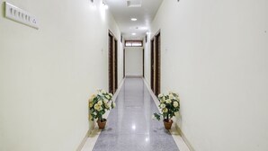 A long, narrow hallway with a tiled floor, white walls, and wooden doors on either side. There are potted plants with white flowers at the hallway's entrance. - Fabhotel Naresh Palace (Raipur)