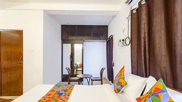 A hotel room with a bed, a small table, two chairs, a wooden door, and a view of a balcony. - Fabexpress Kovalam Residency (Tirupporur)
