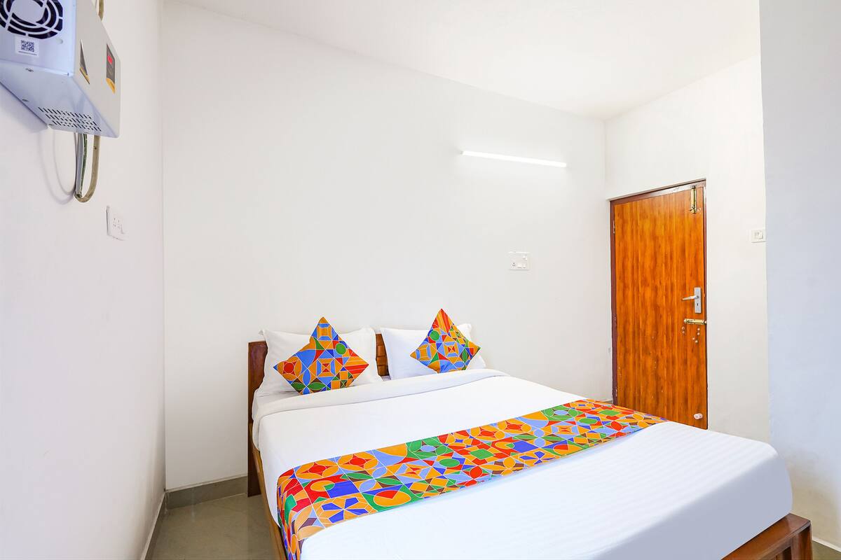A neatly made bed with a colorful geometric-patterned bedspread and pillows, a wooden door, and a wall-mounted air conditioning unit.