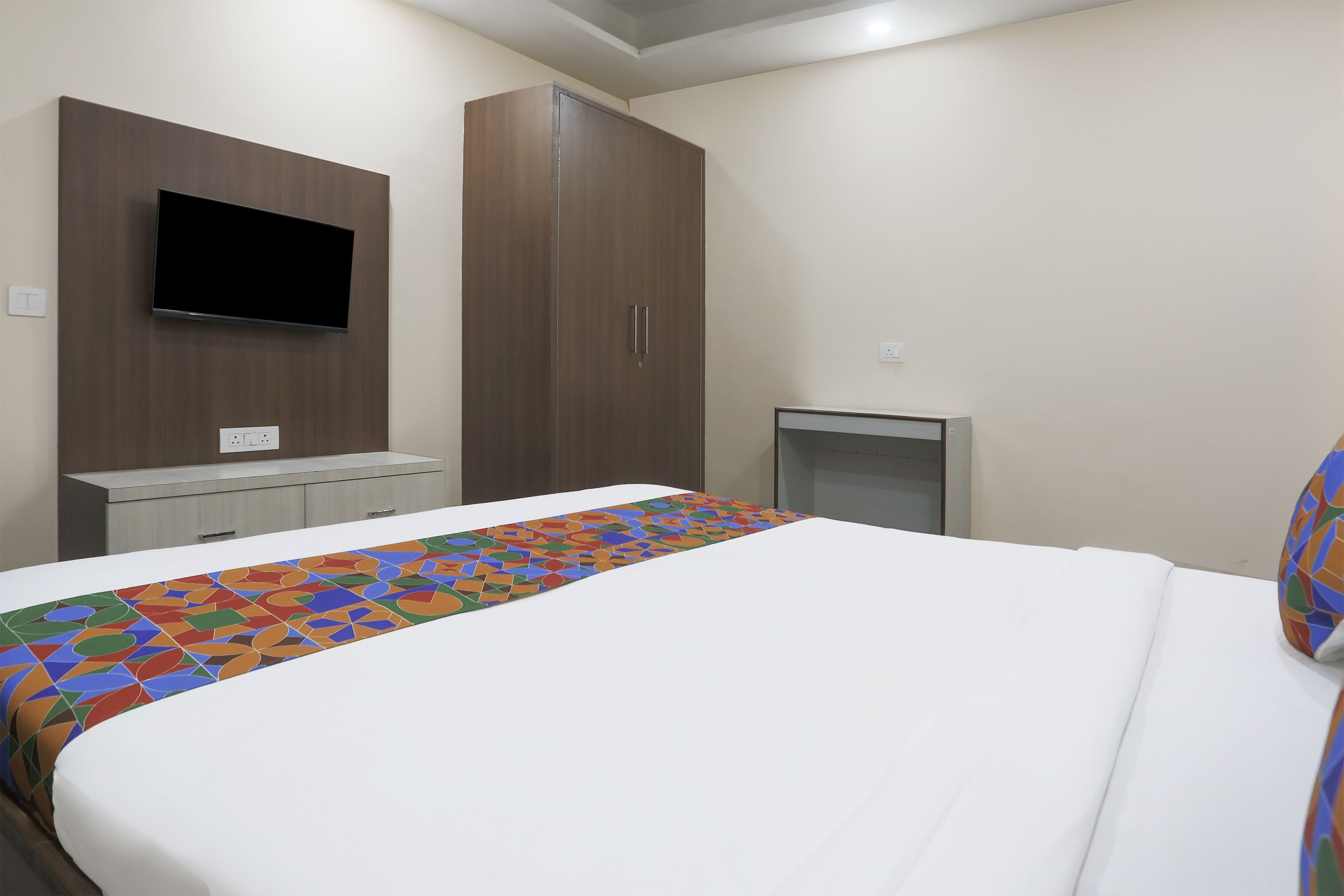 A hotel room with a bed, a TV mounted on the wall, a wardrobe, and a small desk.