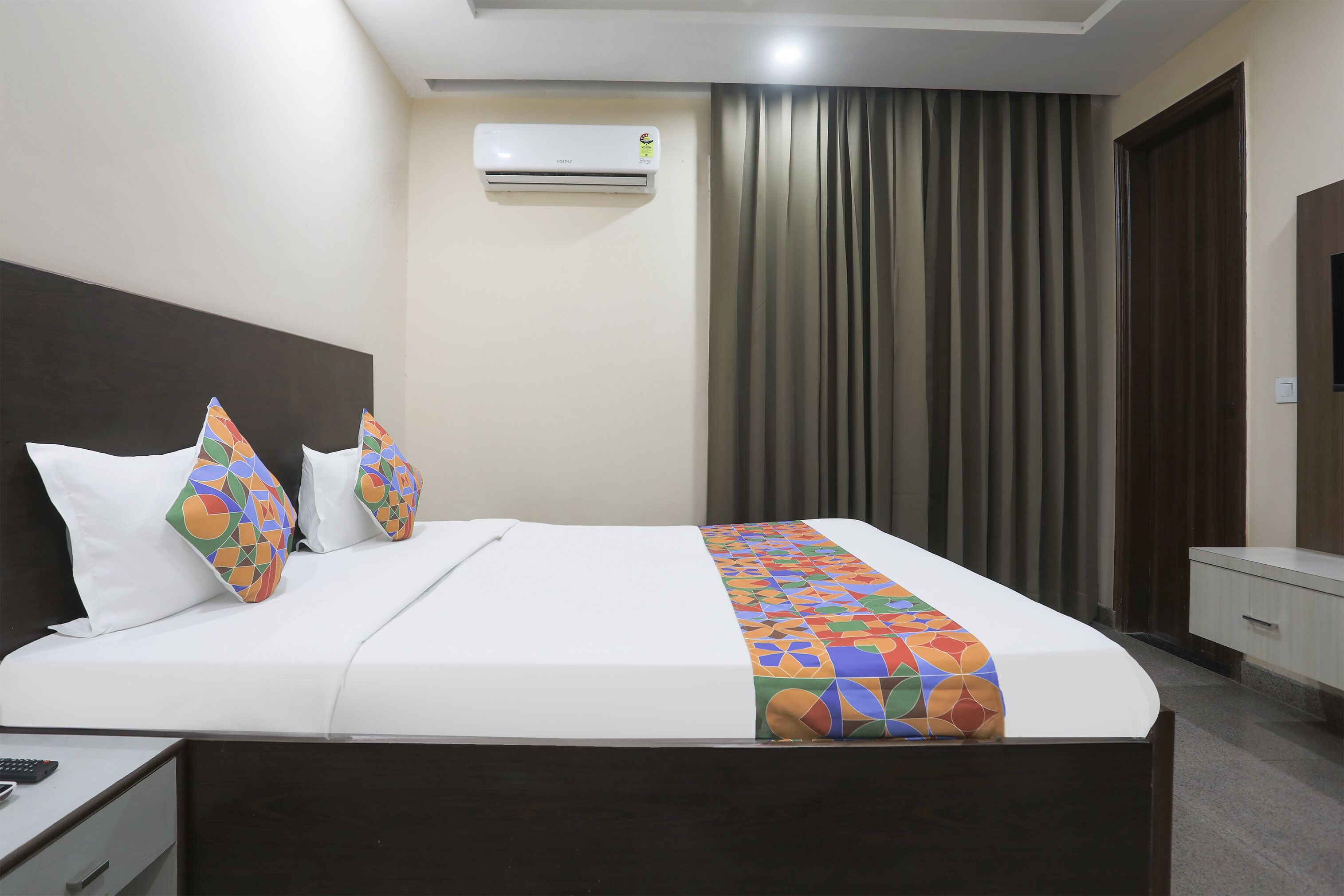A neatly made bed with white bedding and colorful geometric pillows, a bedside table with a remote control, and a wall-mounted air conditioner.