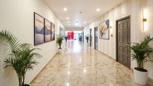 Pasillo de hotel con suelo de mármol, plantas en macetas, cuadros en las paredes y una puerta con una lámpara.