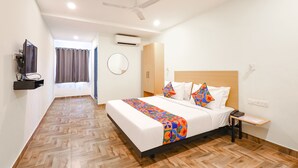 Habitación de hotel con cama, escritorio, silla, ventilador de techo, unidad de aire acondicionado, televisor y ventana con cortinas.